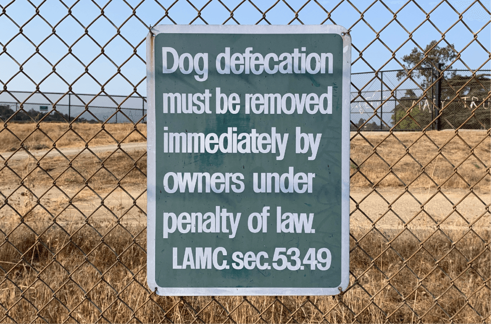 A handpainted sign hanging on a chickenwire fence that reads "Dog defecation must be removed immediately by owners under penalty of law. LAMC. sec. 53.49"