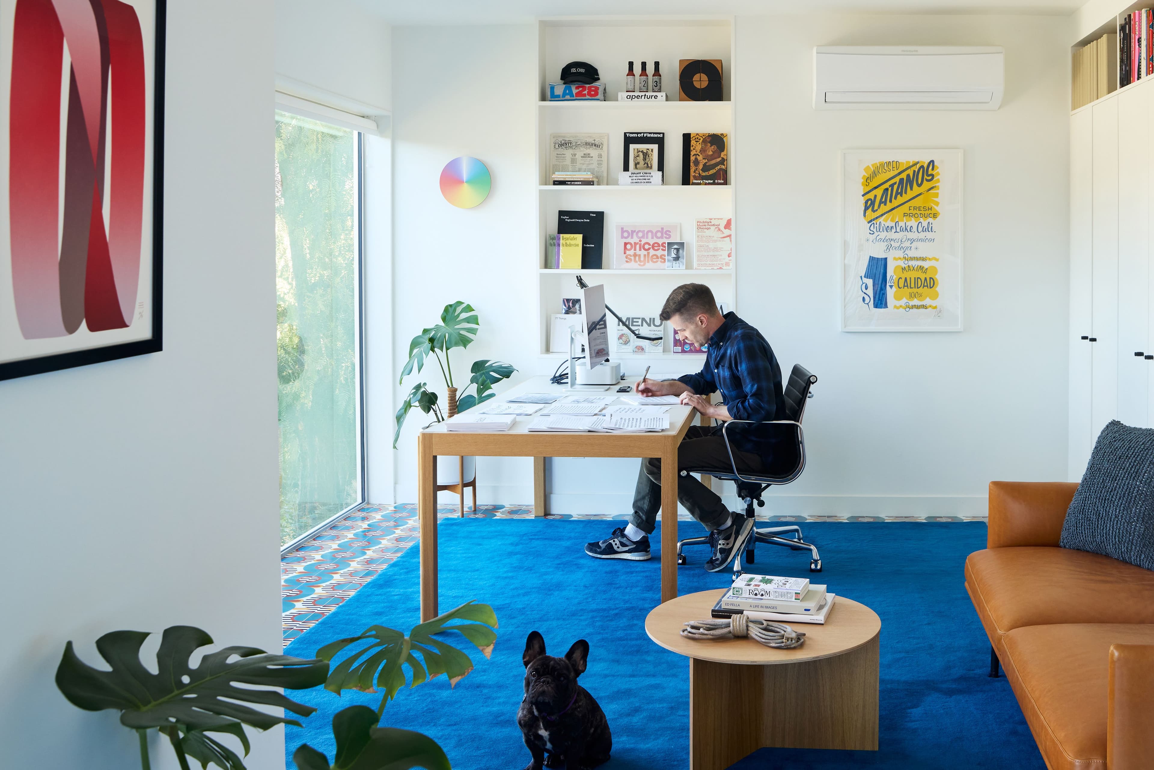 The image shows a modern, bright home office or studio space with clean lines and minimalist decor. The room features a large window allowing natural light to stream in, providing a view of greenery outside. A desk sits near the window, with a sleek monitor and office supplies neatly arranged on it. A comfortable ergonomic office chair is positioned in front of the desk. Behind the desk, built-in white shelves display various design books, objects, and memorabilia, adding a touch of personality to the room. On the wall to the right, there is a framed art piece with sheet music and abstract imagery. The room has a blue rug that contrasts with the light wood furniture and a cozy, stylish couch in the foreground. The overall aesthetic is clean, creative, and well-organized, blending productivity with artistic inspiration.